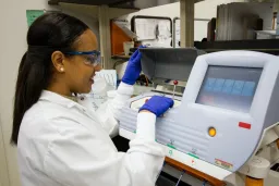 Woman in lab coat, gloves and goggles looking at screen